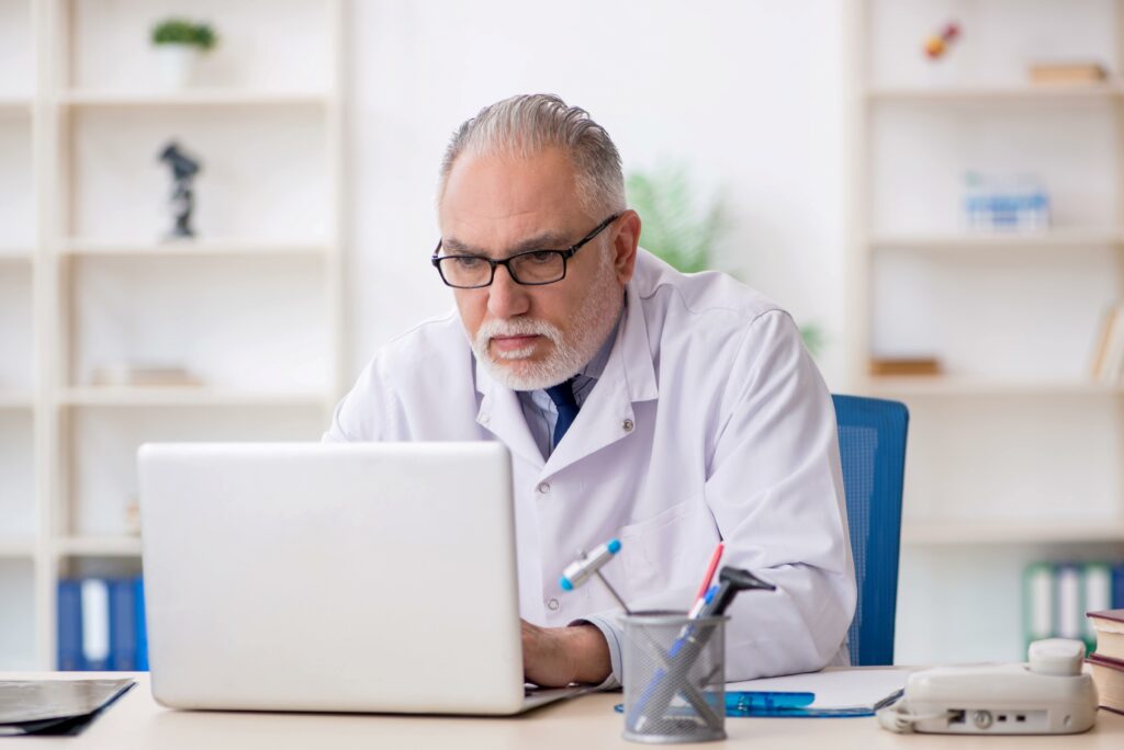 A doctor in black-framed glasses and white medical coat on his computer going over his marketing data and trying to figure out how to grow his practice.